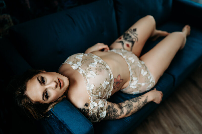 A woman in white lace lingerie lays across a blue couch in a Luxury Boudoir studio