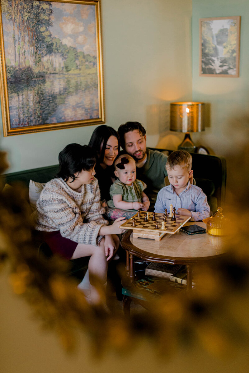 A family of five enjoys quality time in a cozy room. They're gathered around a coffee table with a chessboard. The room features warm lighting, a green wall, and framed artwork. The mood is joyful and relaxed.