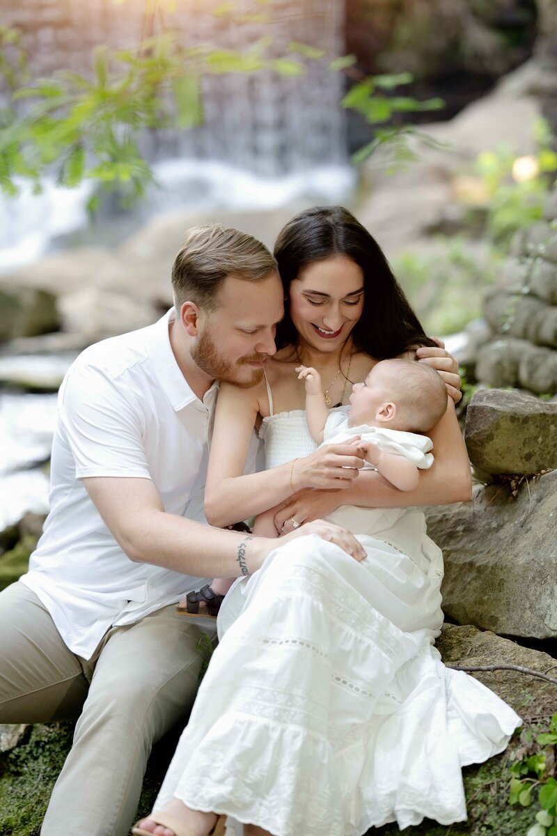 Mom and Dad sitting on a rock by the creek and gazing down lovingly at 4 month old baby boy.