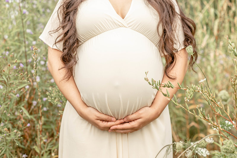 close up of pregnant woman belly with mom holding belly at the bottom with both hands wearing an ivory colored maternity dress.