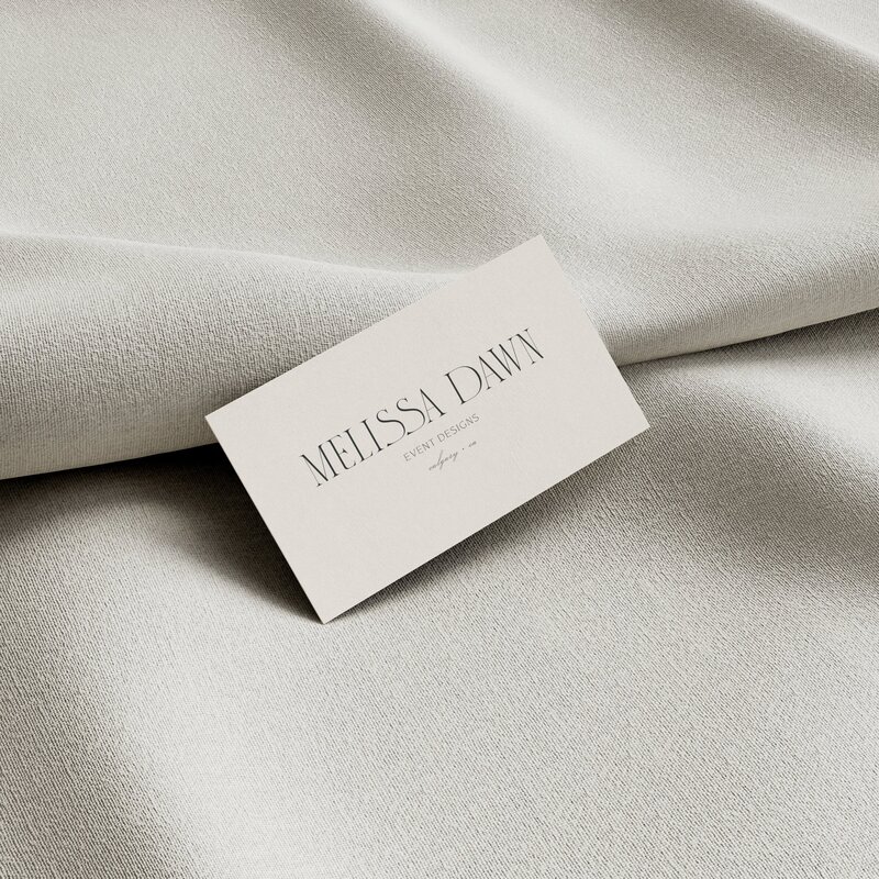 A business card with the name "Melissa Dawn" lies on a textured beige fabric surface, showcasing an elegant wedding planner logo.