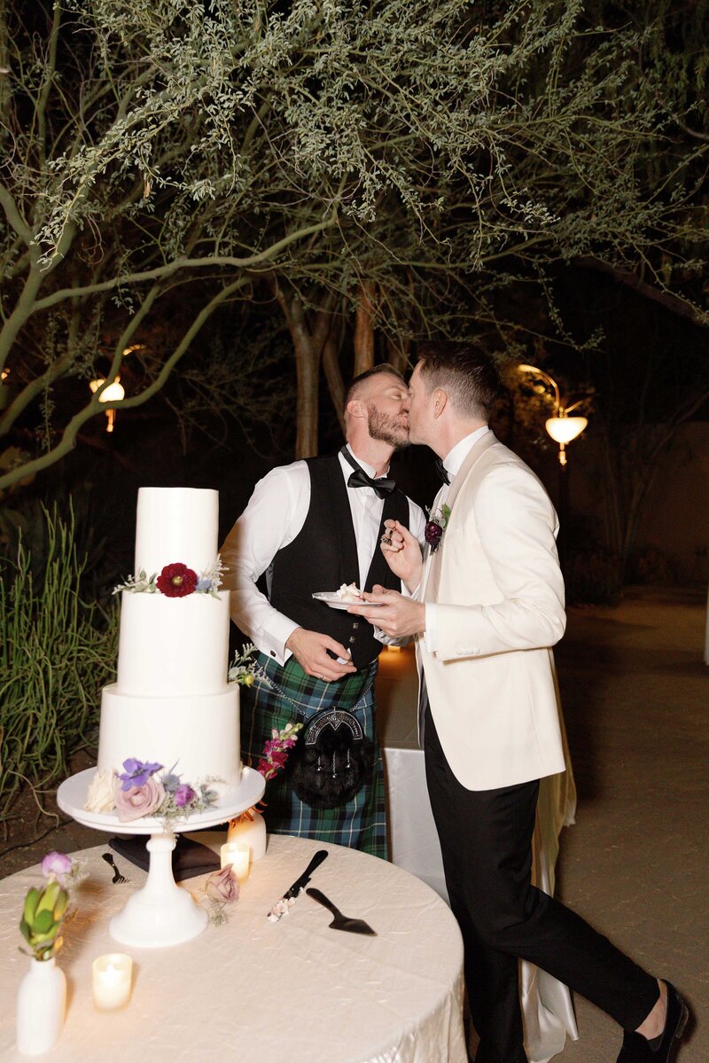 Two grooms kiss after cutting three-tiered wedding cake.