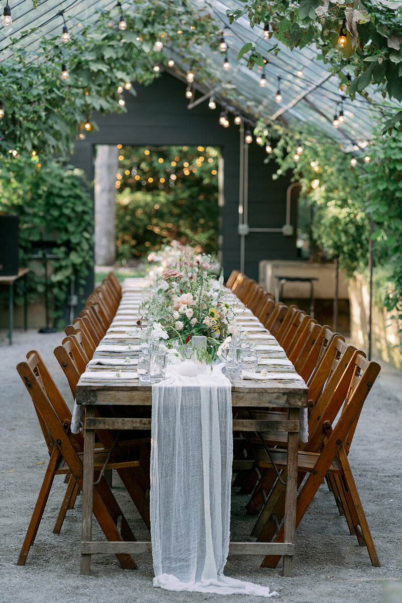 A long reception table decorated with candles and lush florals inside the Glasshouse Community greenhouse during an intimate summer wedding.