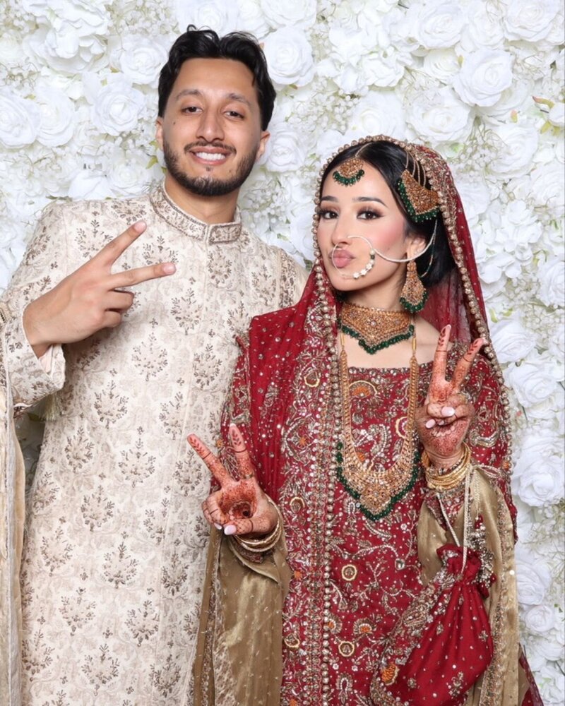 Smiling bride and groom making peace signs and posing playfully at a wedding photo booth with floral backdrop.