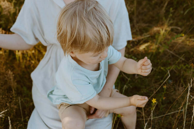 A 3-year old boy sitting at his mother's lap and playing with some hay in his hand at a park in Helsinki in Finland