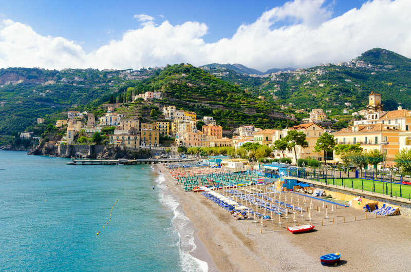 Coastal town with colorful buildings on a hillside overlooks a sandy beach lined with umbrellas and sunbeds. Clear blue water stretches out under a partly cloudy sky, with lush green hills in the background.