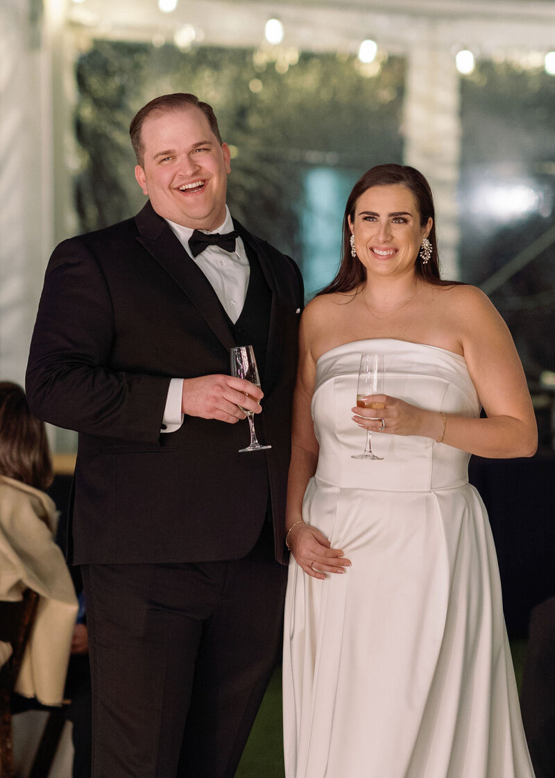 Portrait of groom with classic bowtie tuxedo and bride with soft, wavy ponytail, smiling and standing close, about to kiss, with the soft glow of the sunset behind trees