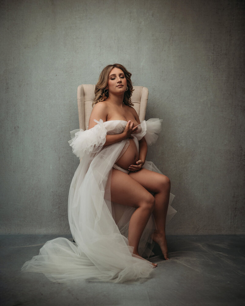 beautiful pregnant woman sitting on beige chair wearing white flowy gown