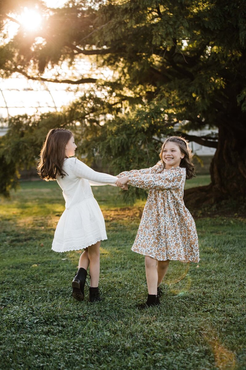 Two young girls holding hands and spinning in a grassy field, smiling and enjoying a sunny day captured by a family photographer in Pittsburgh, PA.