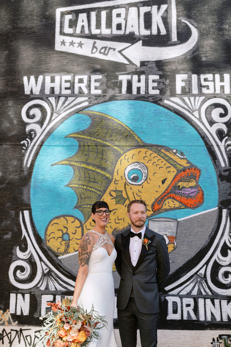 A couple stands in front of a vibrant mural featuring a colorful fish and the text "Where the Fish Drink" at a bar called Callback.