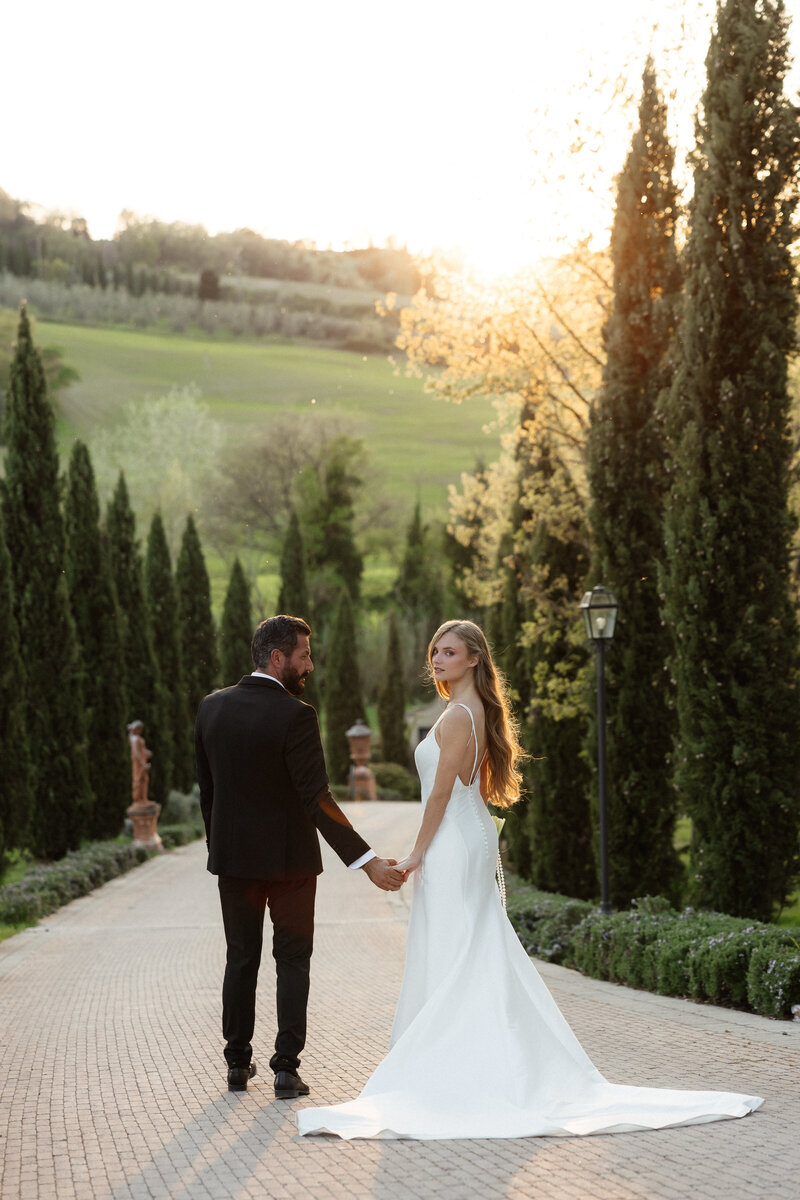 Bride and groom holding hands during sunset in between two rows of Cypress trees in Italy
