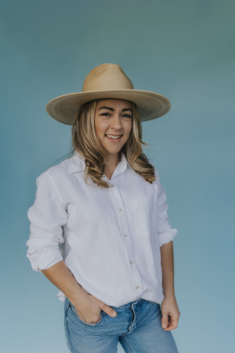 Photo of Alexandra Boynton, owner of Peaches and Poppies Floral, standing in front of a blue wall wearing jeans and a white button down shirt and straw hat smiling at the camera