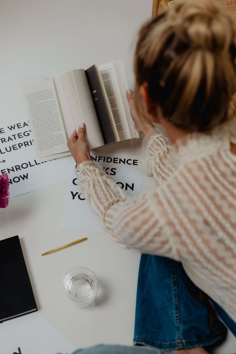 Overhead view of a woman reading a book at a desk surrounded by motivational printouts, a notebook, pen, and glass of water—capturing a calm, focused moment of personal or financial growth.