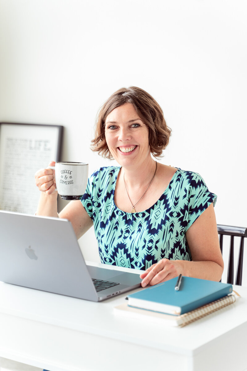 Kate Daniel, a Calgary brand photographer, smiles at her desk with a laptop, holding a “Coffee & Cameras” mug. A bright, inviting, and professional brand photo capturing a relaxed and creative work setting.