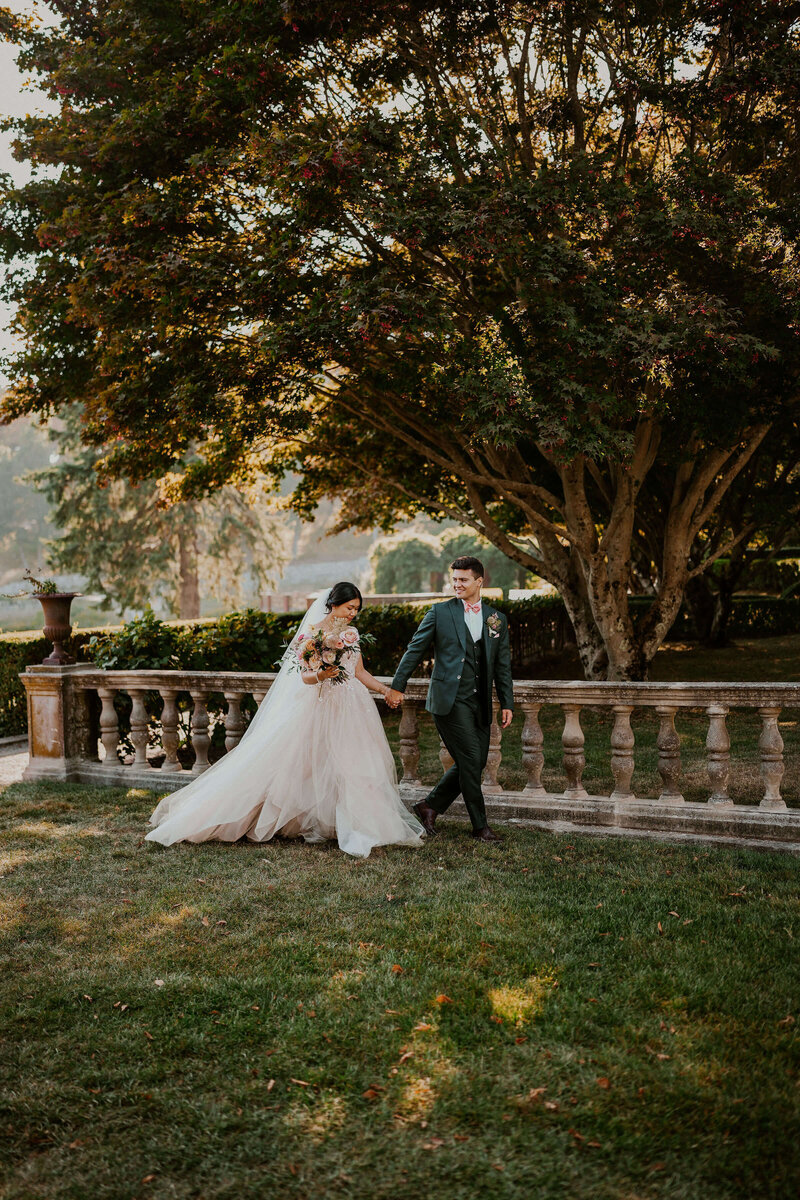 Bride holding a bouquet while walking through a garden pathway hand in hand with groom, captured in a candid moment with natural sunlight and trailing gown.