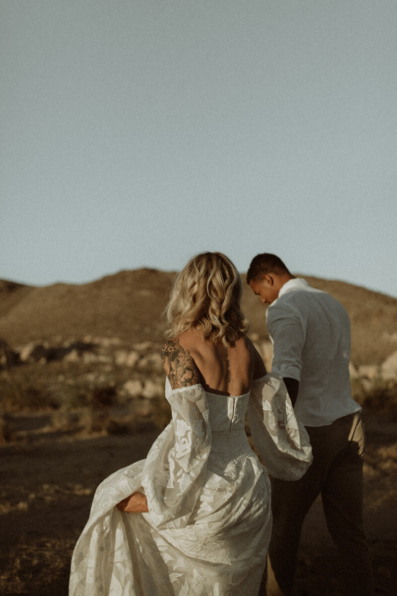 Couple eloping in Joshua Tree National Park at sunset. Walking away from the camera while the sun glistens on their skin and the bride's blonde hair.