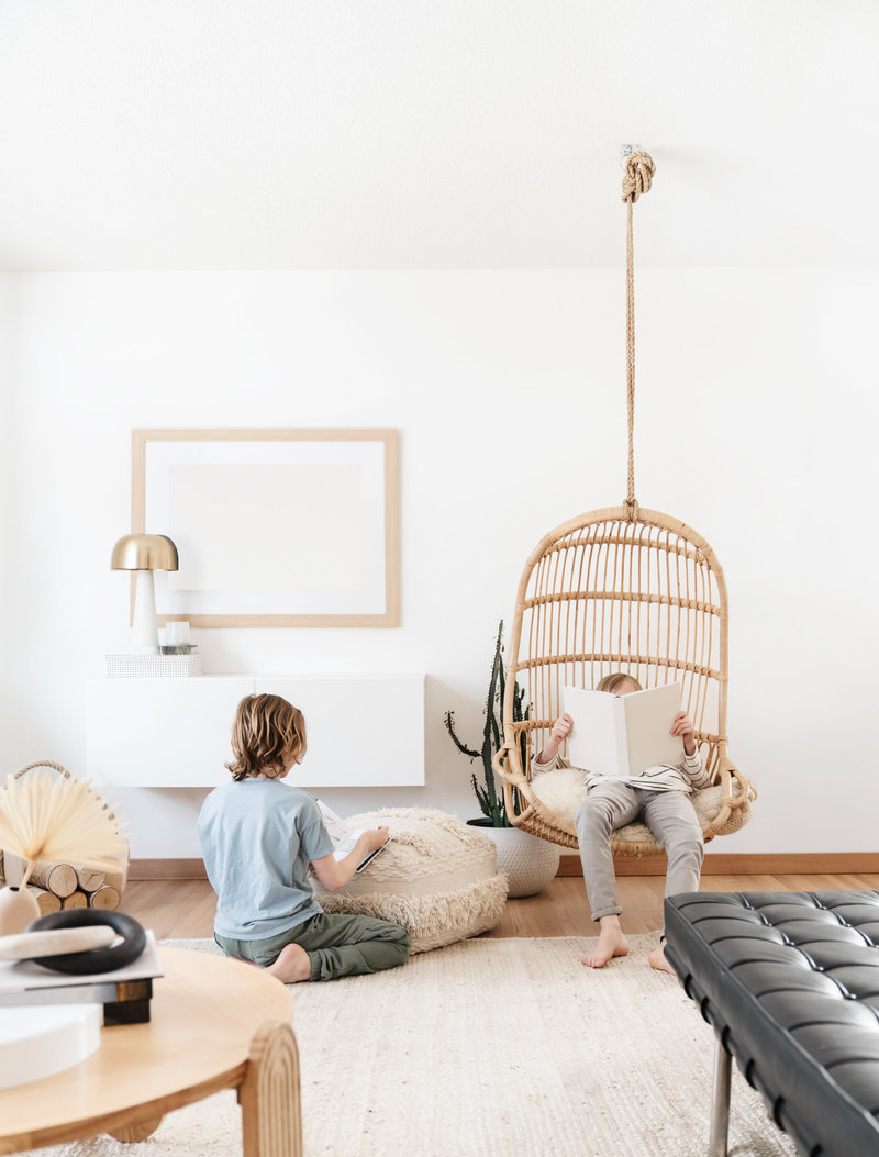 Two children read quietly in a bright and minimal living room, one sitting on the floor and the other relaxing in a hanging wicker chair – Family Autism Support Coach at Chantal Hewitt Coaching