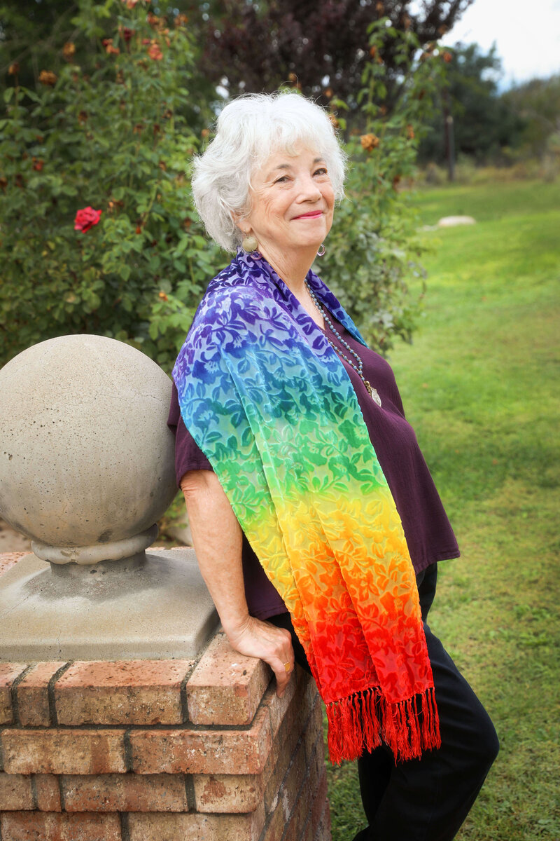 Side profile of  Dr. Samarah Grace wearing a rainbow shaw leaning on brick pillar smiling at camera with pink rose bush behind her