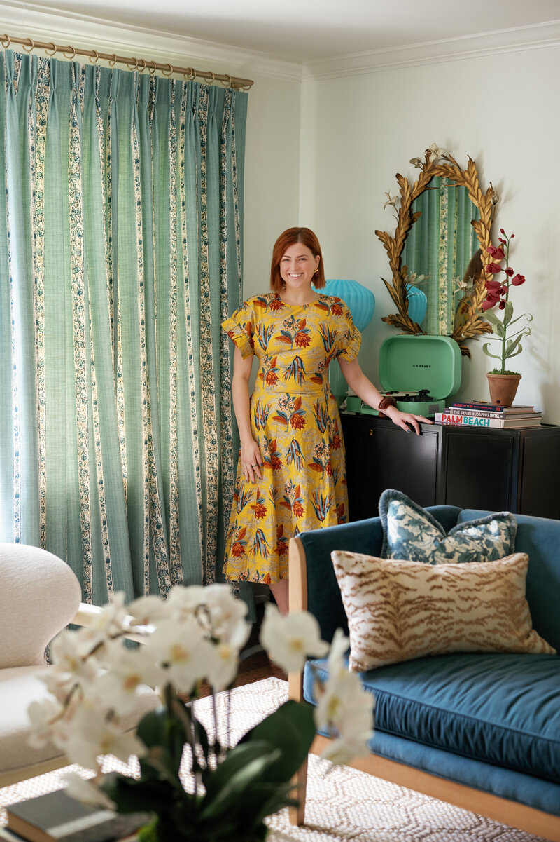 Chelsey Heslop in a yellow pattern dress standing in a living room with her arm on a black console table next to blue pattern curtains