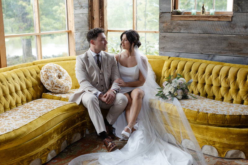Bride and groom sit next to each other on a yellow couch while gazing into each other's eyes