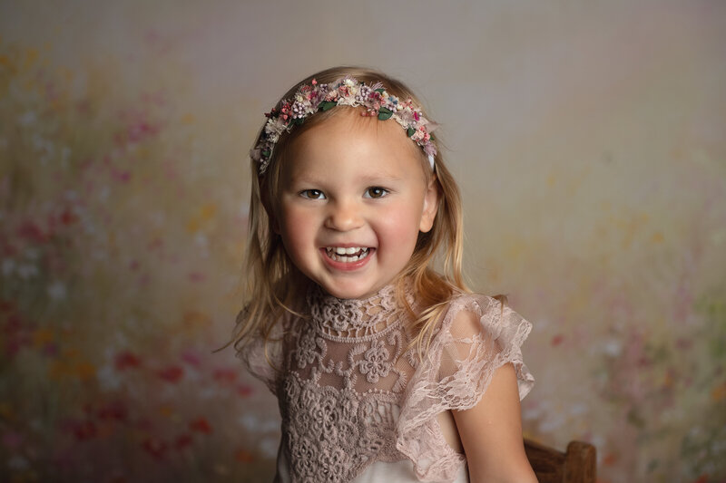A toddler girl smiles big in a pink dress in a studio