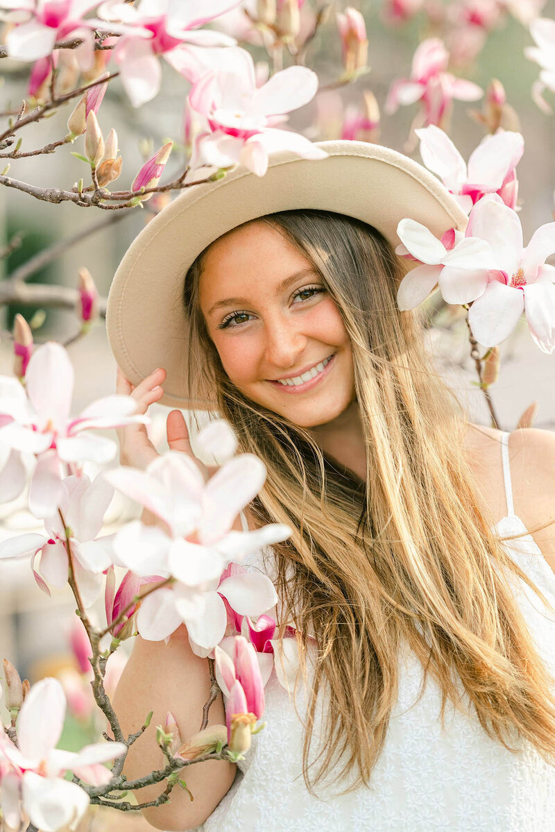 Girl wearing a tan hat and white dress in front of pink and white magnolia flowers