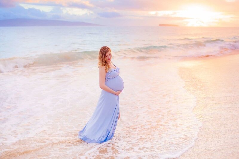 Brunette in blue Kasia Kulenty maternity gown cradles her pregnant belly during Love + Water maternity session on Maui