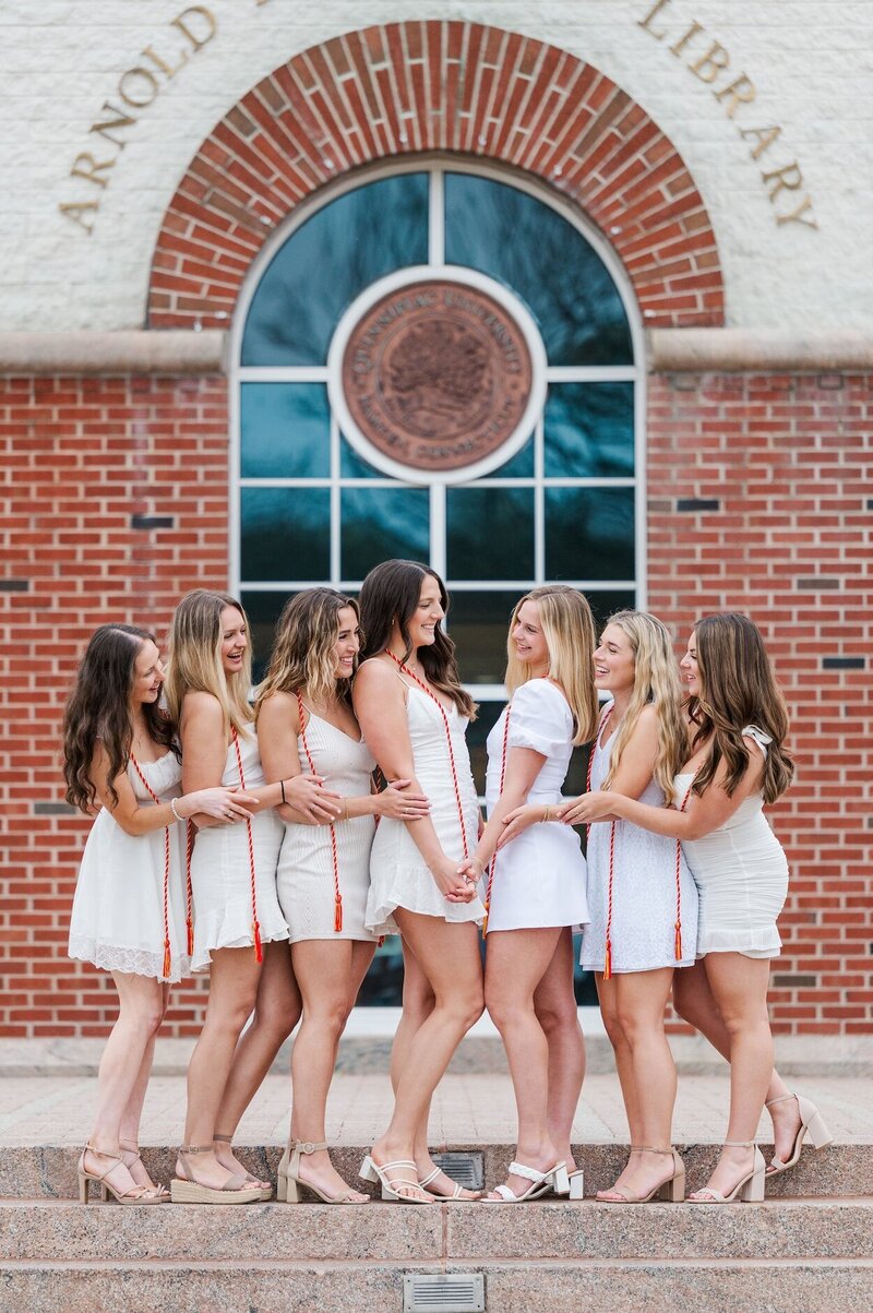 A group of seven Quinnipiac graduates hold hands with one another and smile at each other in front of Arnold Bernhard Library.