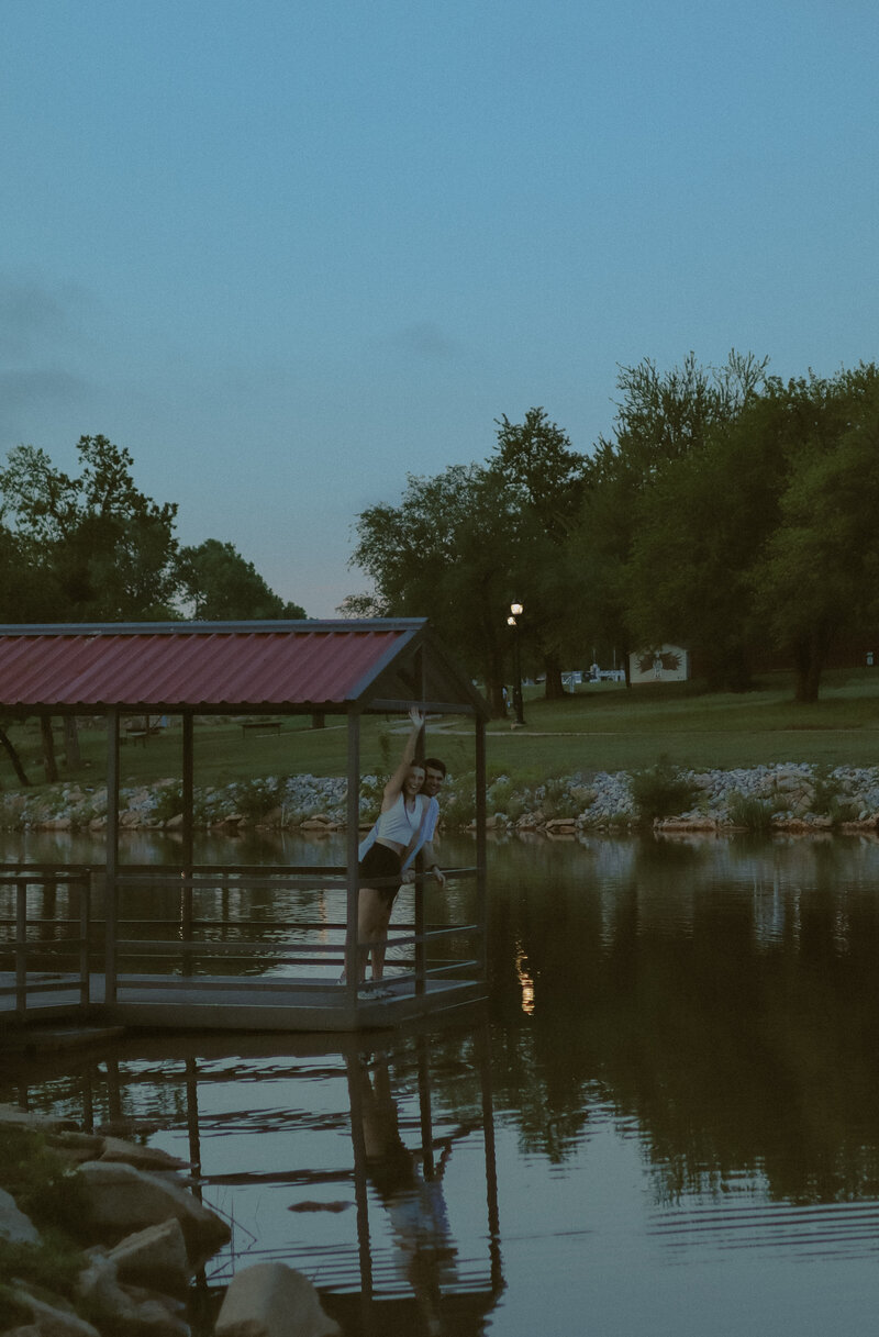 A couple can be seen on a dock waving at the camera from a distance away. They are surrounded by water and trees.