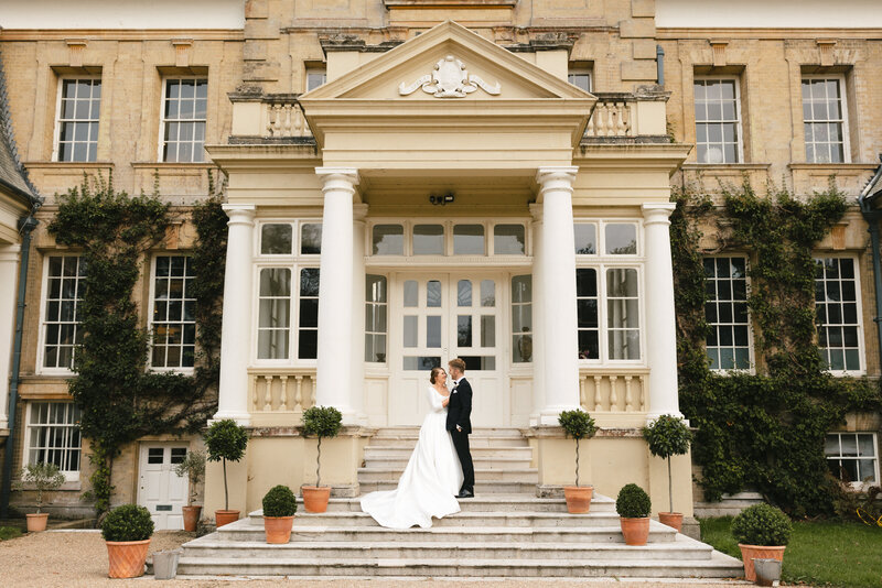 A couple in wedding attire stands on stone steps outside Pylewell Park, an ivy  covered. building with large windows and columns. She wears a white gown, and he is in a black suit. Potted plants line the steps.