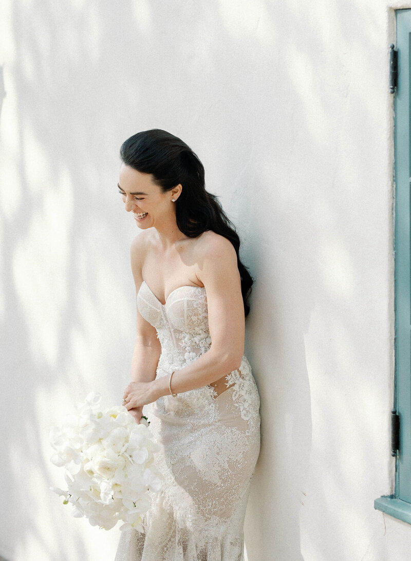 A fair-skinned bride in a lacy strapless wedding gown leans against a white exterior wall, laughing and holding a bouquet of white flowers