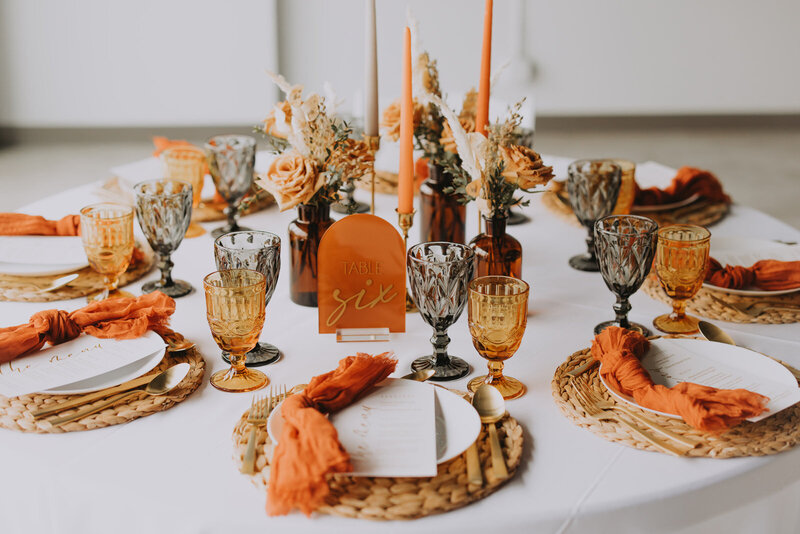 Round wedding table styled with amber glassware, woven placemats, burnt orange napkins, terracotta candles, and dried floral centerpieces for a rustic autumn-inspired reception.