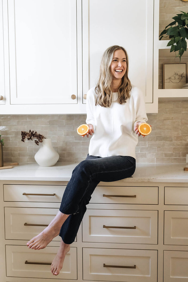 Erica Taylor smiling in the kitchen holding orange halves—encouraging moms to feel energized, nourished, and confident in their wellness