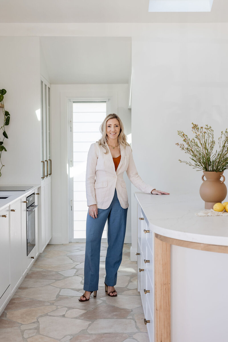 A woman in a white blazer stands in a modern kitchen, smiling, with a vase and lemons on the counter. Her elegant space is the work of a Newcastle Kitchen and Bathroom Designer renowned for blending functionality with style.