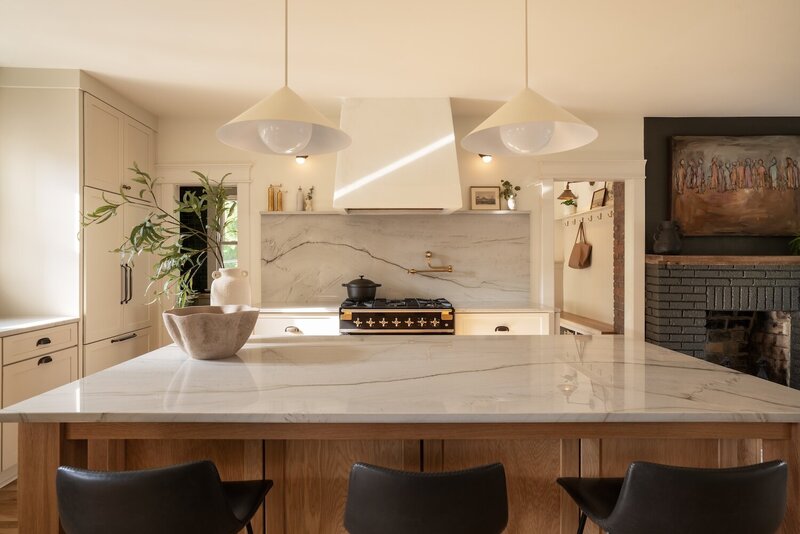 Gourmet chef kitchen with custom black and gold oven, white modern range hood, large marble backsplash, massive island with counter seating, and natural light from backyard windows.
