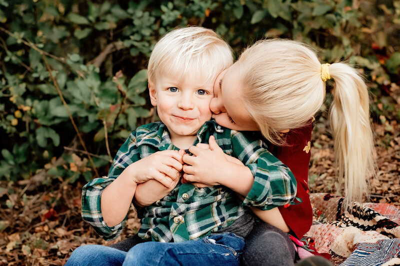 up close portrait of a toddler boy sitting on his older sister's lap.  She has her arms around him and he has his hands placed over hers.  She is leaning over and kissing his cheek and he has a smirk on his face.