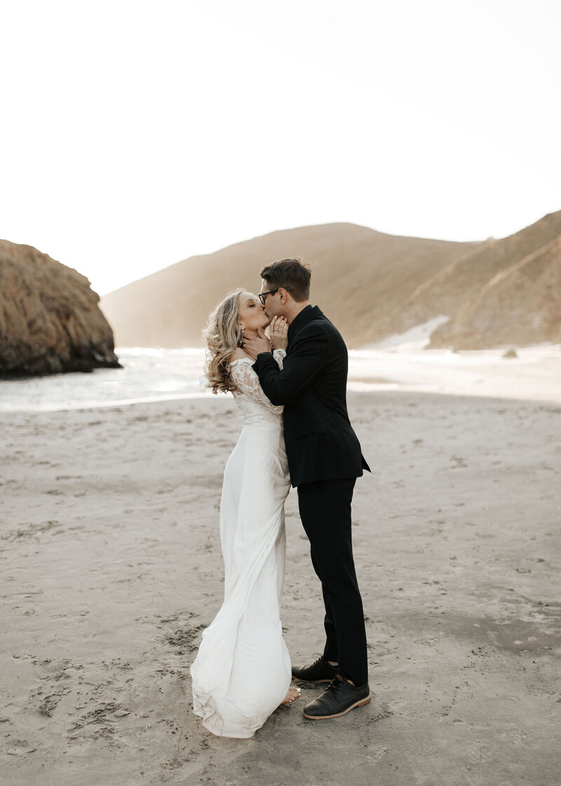 bride and groom standing and kissing in front of beach mountains
