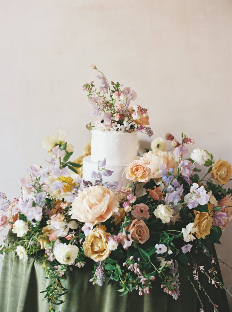 white wedding cake with peach, yellow, and purple flowers and a dark green tablecloth