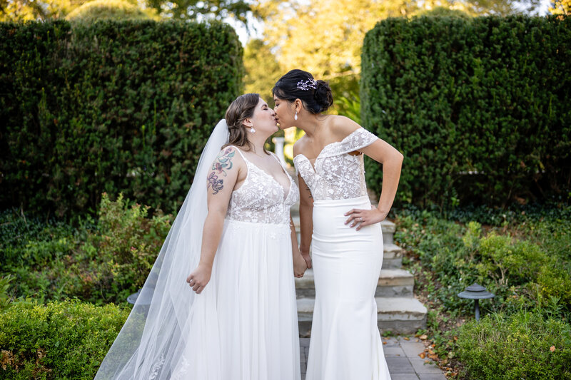 laotian bride in off the shoulder white gown with a curvy white bride in a spaghetti-strap gown holding hands and kissing each other on a stone path in green hedge garden