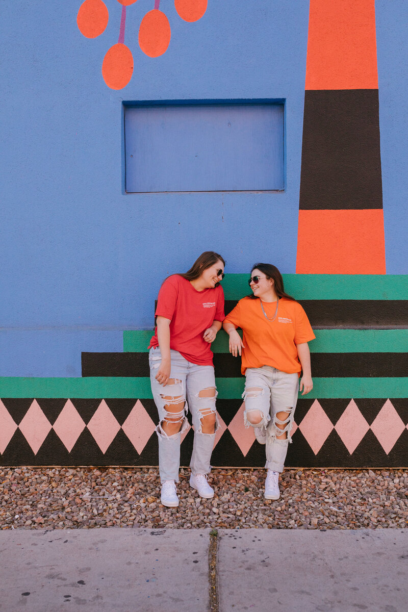 Two girls leaning up against a colorful wall looking at each other and smiling.