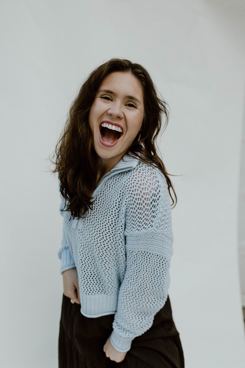 A woman laughing at the camera standing in front of a white backdrop. Wearing a light blue sweater and a brown skirt.