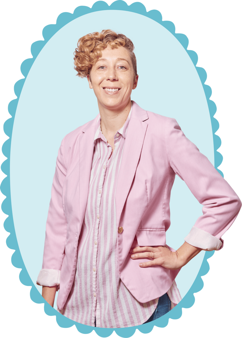 Megan Kranzler, Owner, standing and smiling with her left hand on her hip, wearing a button up shirt featuring pink and white vertical stripes under a pink blazer with the sleeves rolled up