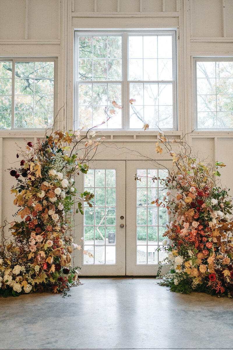 Large fall wedding floral arch installation featuring lush seasonal blooms, including roses, dahlias, chrysanthemums, and autumn foliage in warm tones of peach, burgundy, gold, and ivory, arranged symmetrically around white double doors in an indoor ceremony space—luxury garden-style floral design.