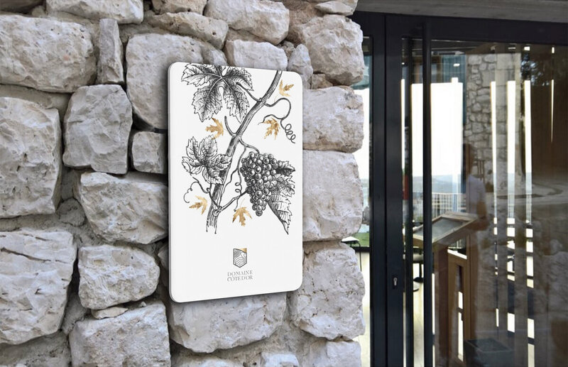 Panneau d'entrée du domaine Côte d'Or posé sur un mur de pierres brutes avec illustration d'une branche de vigne et feuilles d'or, crée par le Studio Youmé.