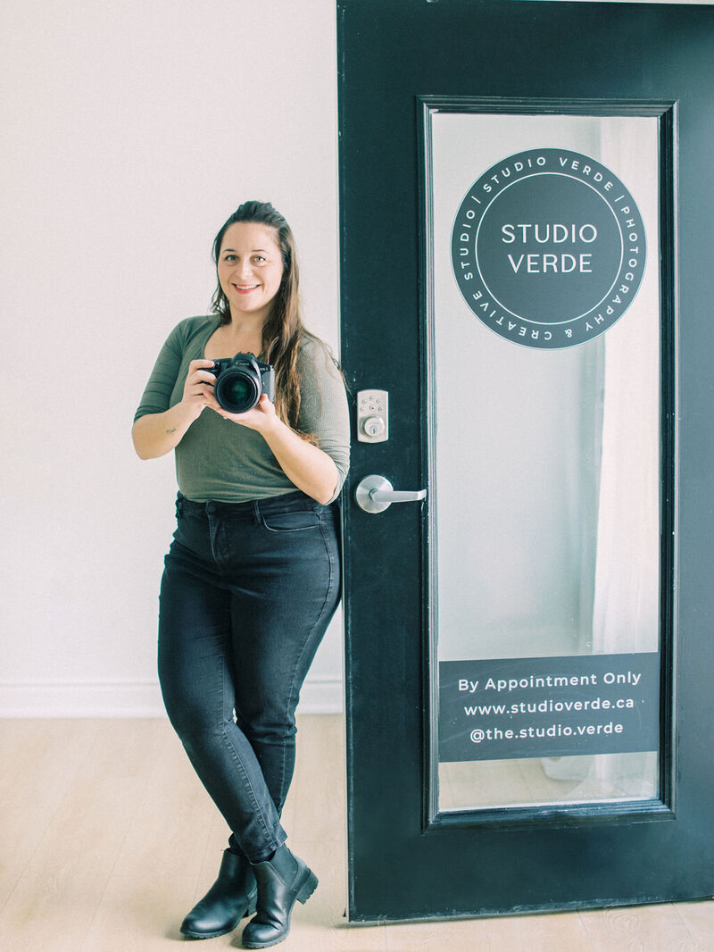 Sara holding her camera wearing an olive green shirt and black pants standing in front of her Studio door with a sign that says Studio Verde, By appointment only.