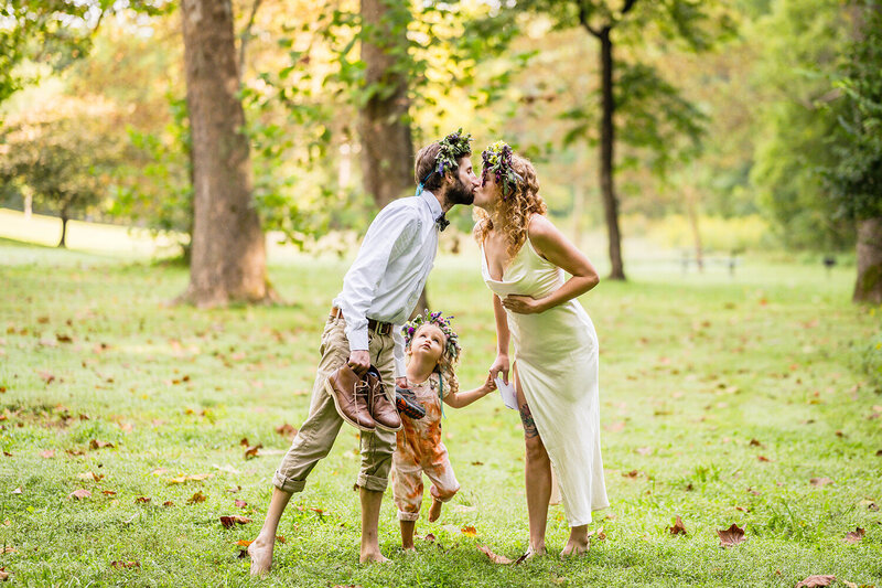 A couple holding their child's hands go in for a kiss while their child looks up curiously during their elopement day at Fishburn Park in Roanoke, Virginia.