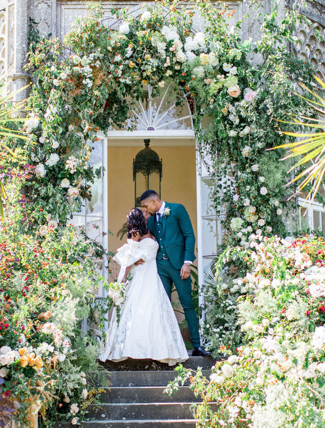 A black bride and groom sharing a kiss standing under an enormous flower arch by All For Love London on their wedding day at Sezincote House with By Chenai