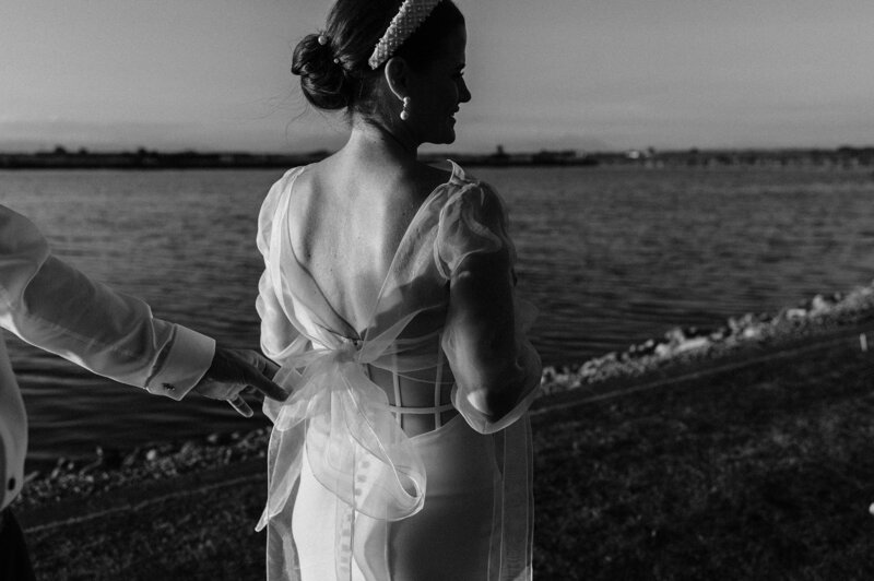 black and white photo of bride in hera couture sheer top with trish peng dress beside a lake at her backyard christchurch wedding