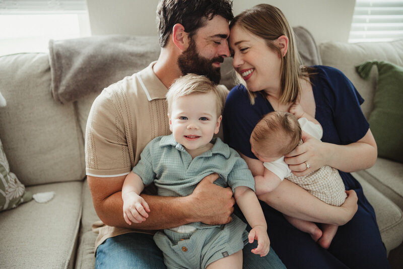 A family holding their newborn son and their toddler while the boy looks directly into the camera smiling during an in-home lifestyle newborn portrait session in Farmington, Missouri. 