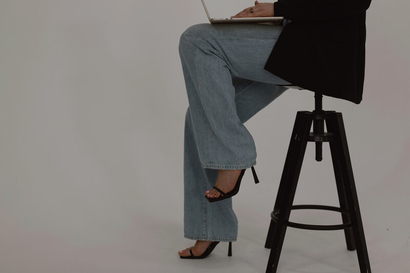 Maria Rose Esposito sitting on a black industrial stool, working on a laptop, wearing light-wash jeans, black blazer, and strappy black heels. The modern, minimal backdrop reflects Sable & Rose’s balance of professionalism and creativity—representing the brand’s collaborative approach to building client-focused PR strategies.